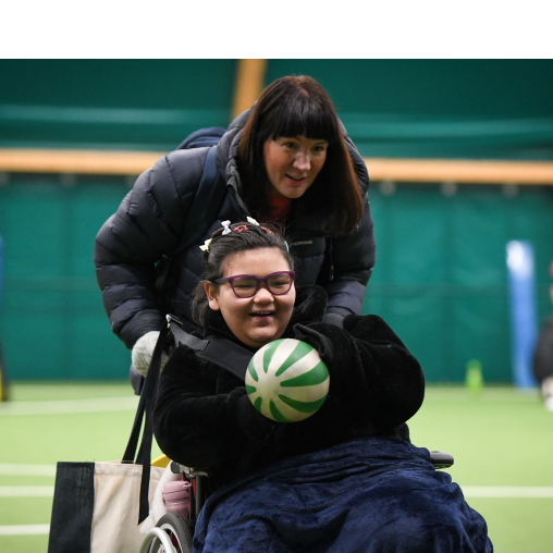 Young girl in a wheelchair holding a ball is being pushed by an adult. 