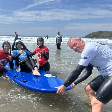 A young disabled person sits on an adapted surfboard in the sea being helped by a group of people.