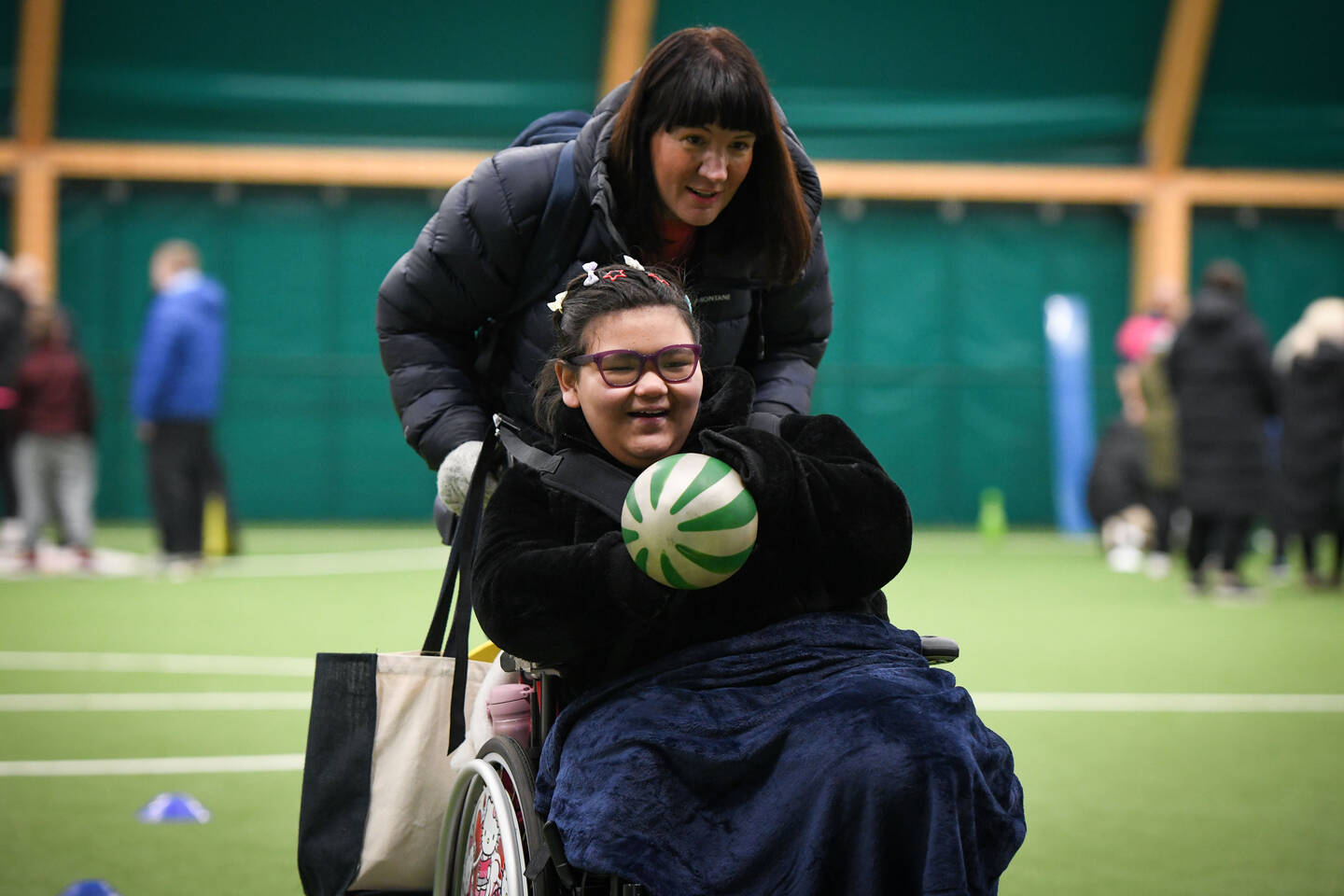 Young disabled girl with a ball is being pushed in a wheelchair by an adult. They about both smiling and chatting.