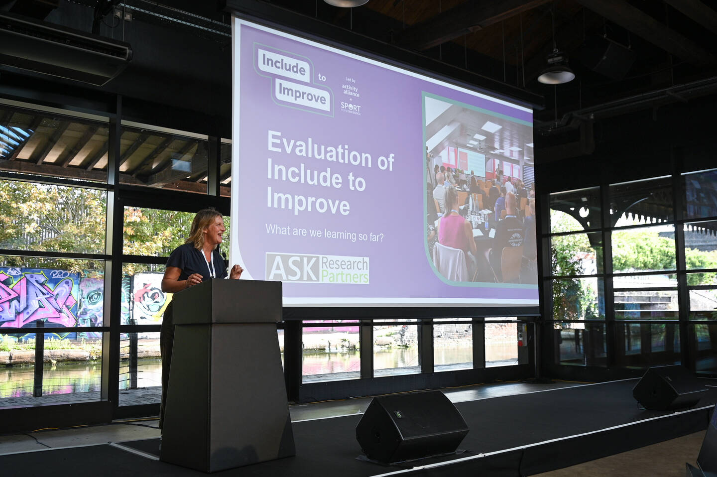 A woman gives a presentation on stage at the I2I celebration event. She is stood in front of a screen.